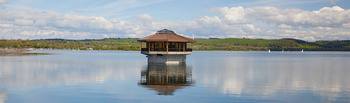 This landscape photograph features the Carsington Water Drain House situated centrally on the Carsington Water reservoir. Taken during a spring morning, the image shows the drain house reflected clearly in the calm lake waters. Surrounding the reservoir, gentle hills and green fields typical of the Peak District can be observed, with wind turbines visible on the distant ridges under a partly cloudy sky. White sailing boats are scattered across the surface of the lake, contributing to the serene scene. The photograph captures both the architectural structure of the drain house and the natural beauty of the location, highlighting Carsington Water as a significant feature in the region.